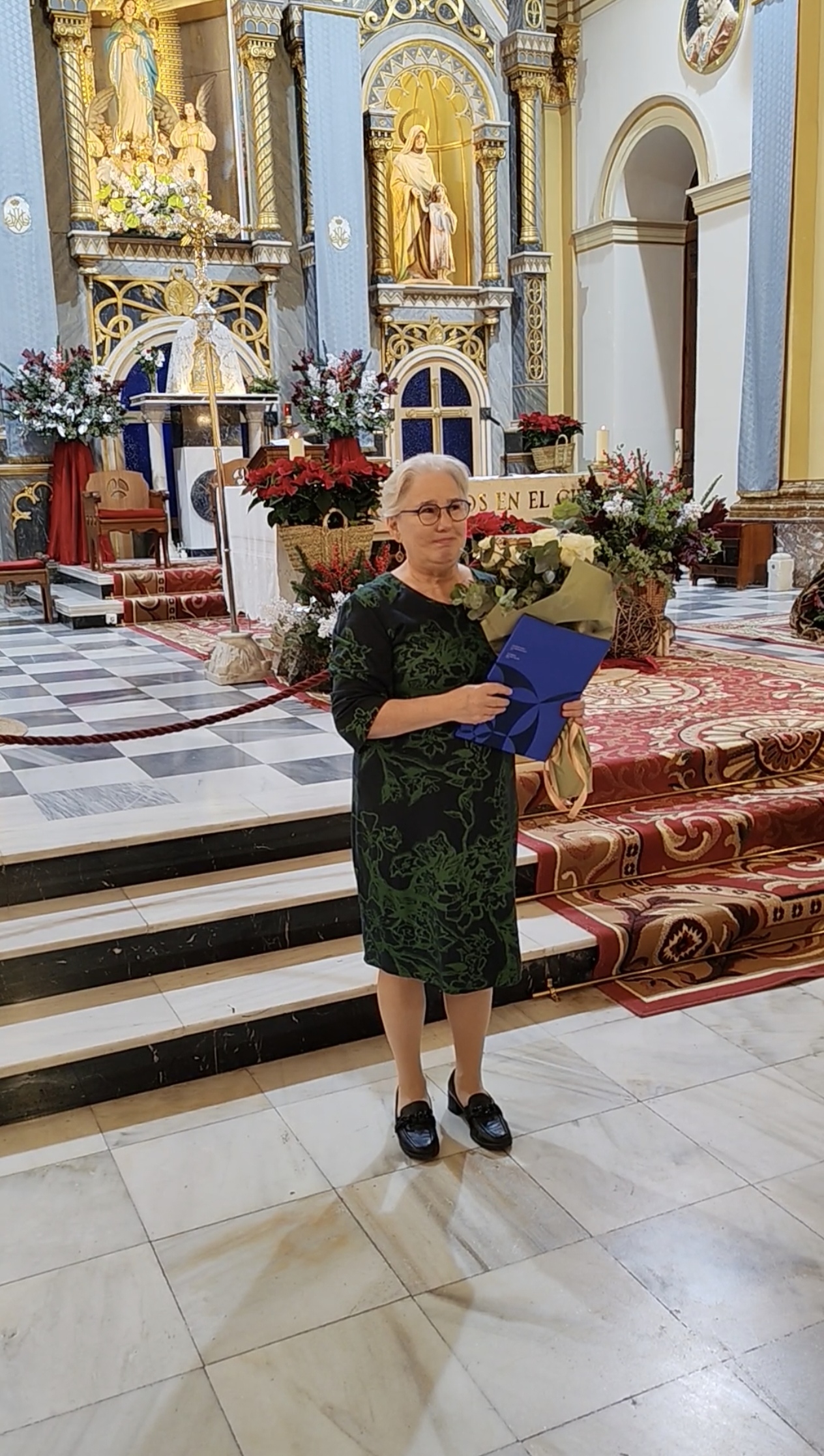 Kuvassa harmaatukkainen, silmälasipäinen nainen sinisessä asussa seisoo alttarin edessä kädessään valkoisia...