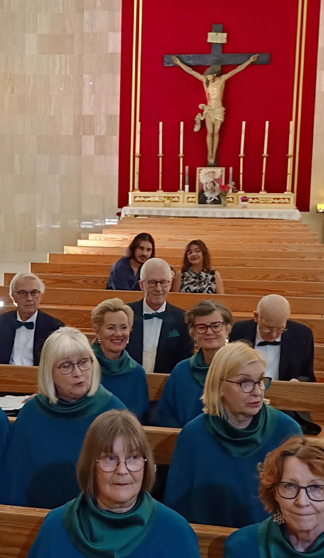 Kuvassa taustalla Kristus ristillä punaista seinävaatetta vasten. Etualalla siniasuisia naisia sekä tummapu...
