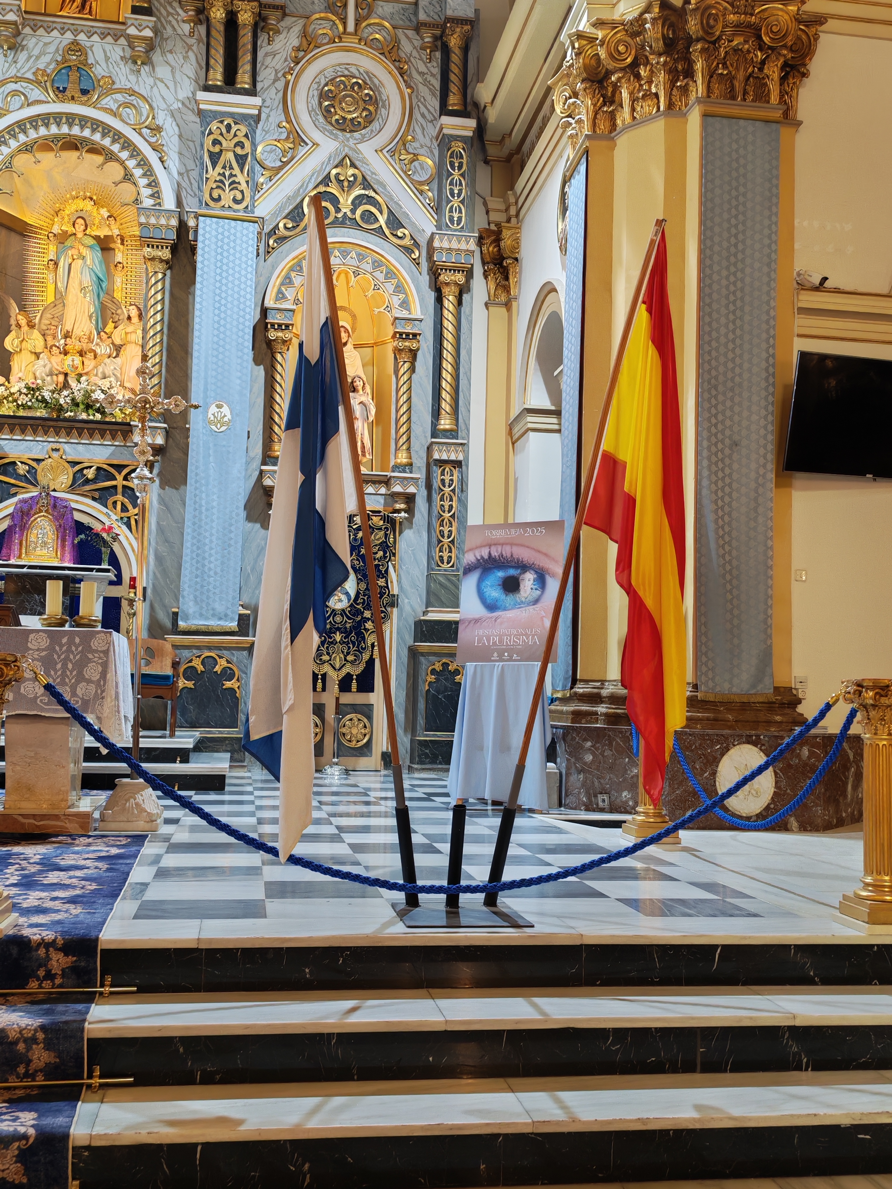 Kuvassa koristeellisessa kirkossa alttarin edessä Suomen ja Espanjan liput. Niiden keskellä näkyy sininen s...