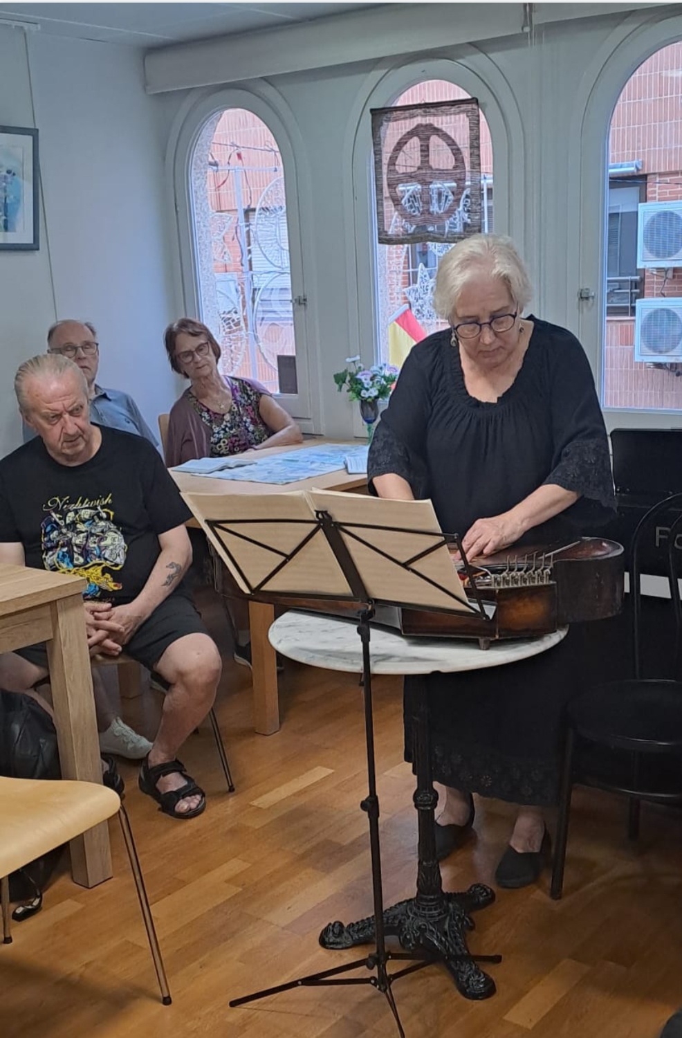 Kuvassa tummapukuinen ja harmaatukkainen nainen soittaa kannelta. Taustalla näkyy yleisöä.