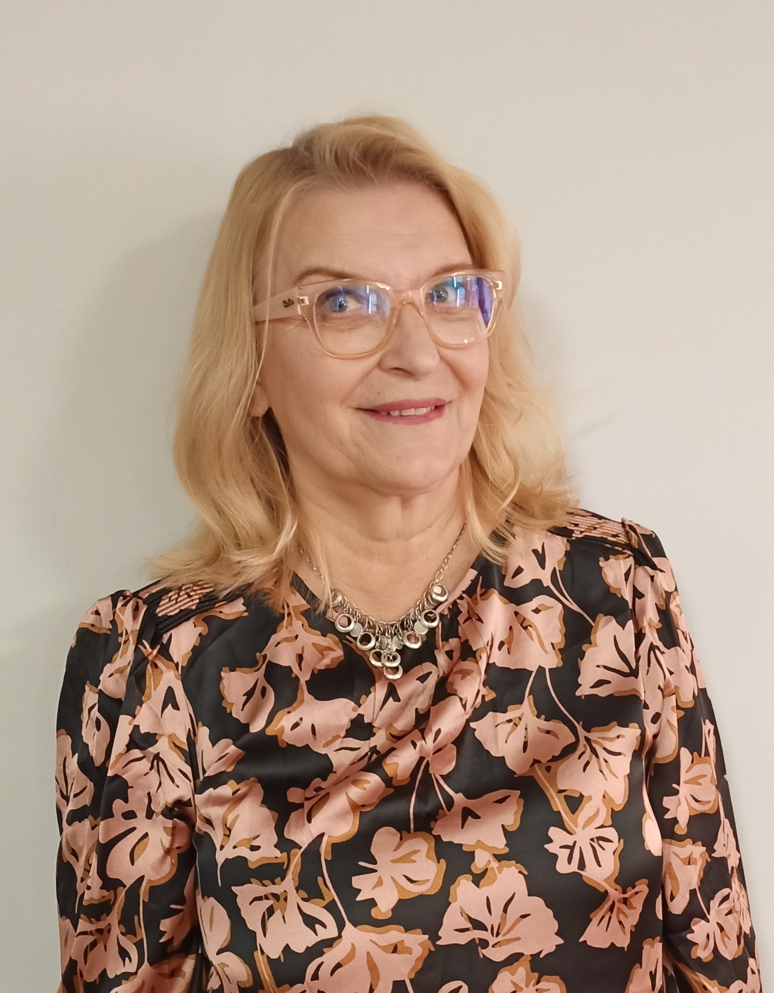 Kuvassa vaaleatukkainen, silmälasipäinen nainen hymyilee kukikkaassa puserossa.